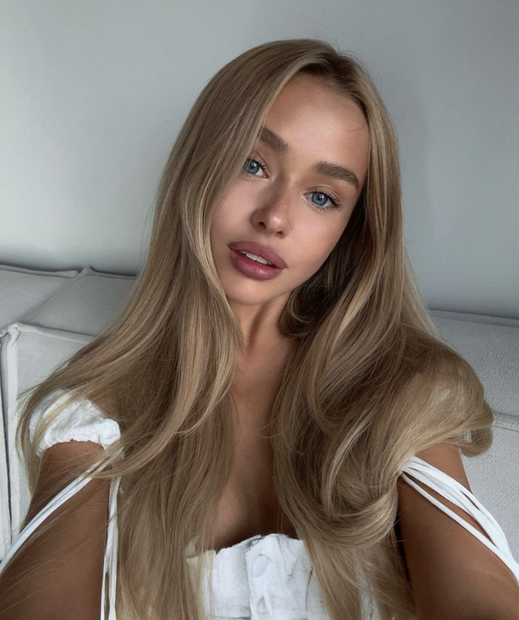Nicole sits on a light couch in a white off-the-shoulder top with thin straps, looking at the camera.