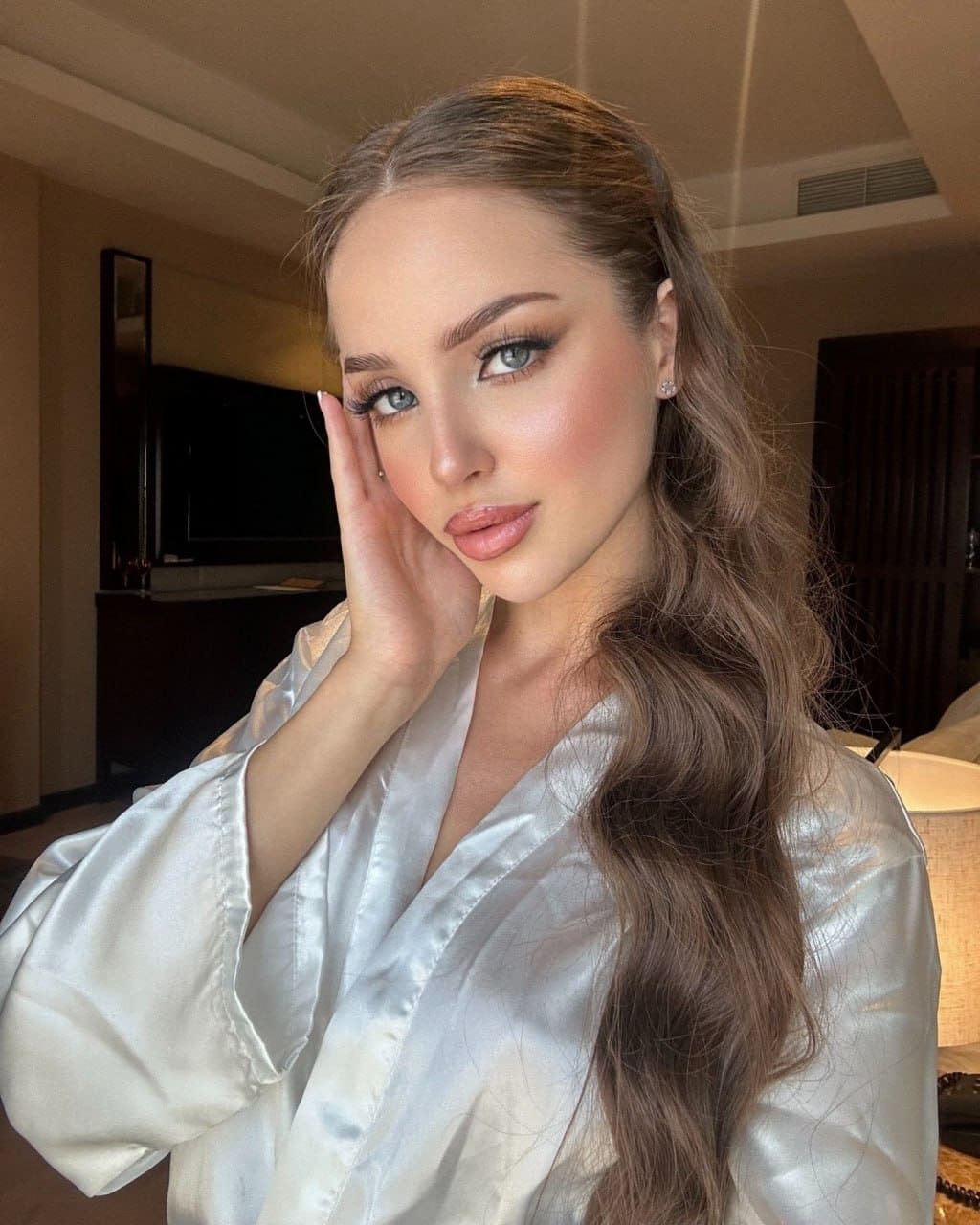 A woman in a loose white satin robe rests her hand against her cheek in a room with dark wood furniture and a television.
