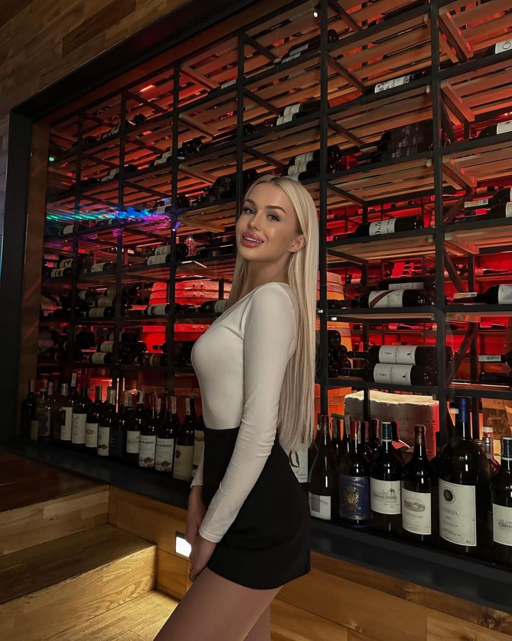 A blonde woman stands in profile before wine racks, wearing a white long-sleeved top and short black skirt.