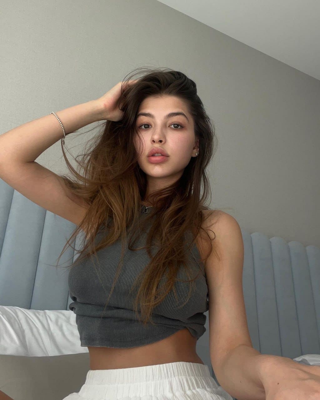 A woman sits on a bed with a blue headboard, wearing a grey ribbed crop top and white pleated bottoms, touching her hair.