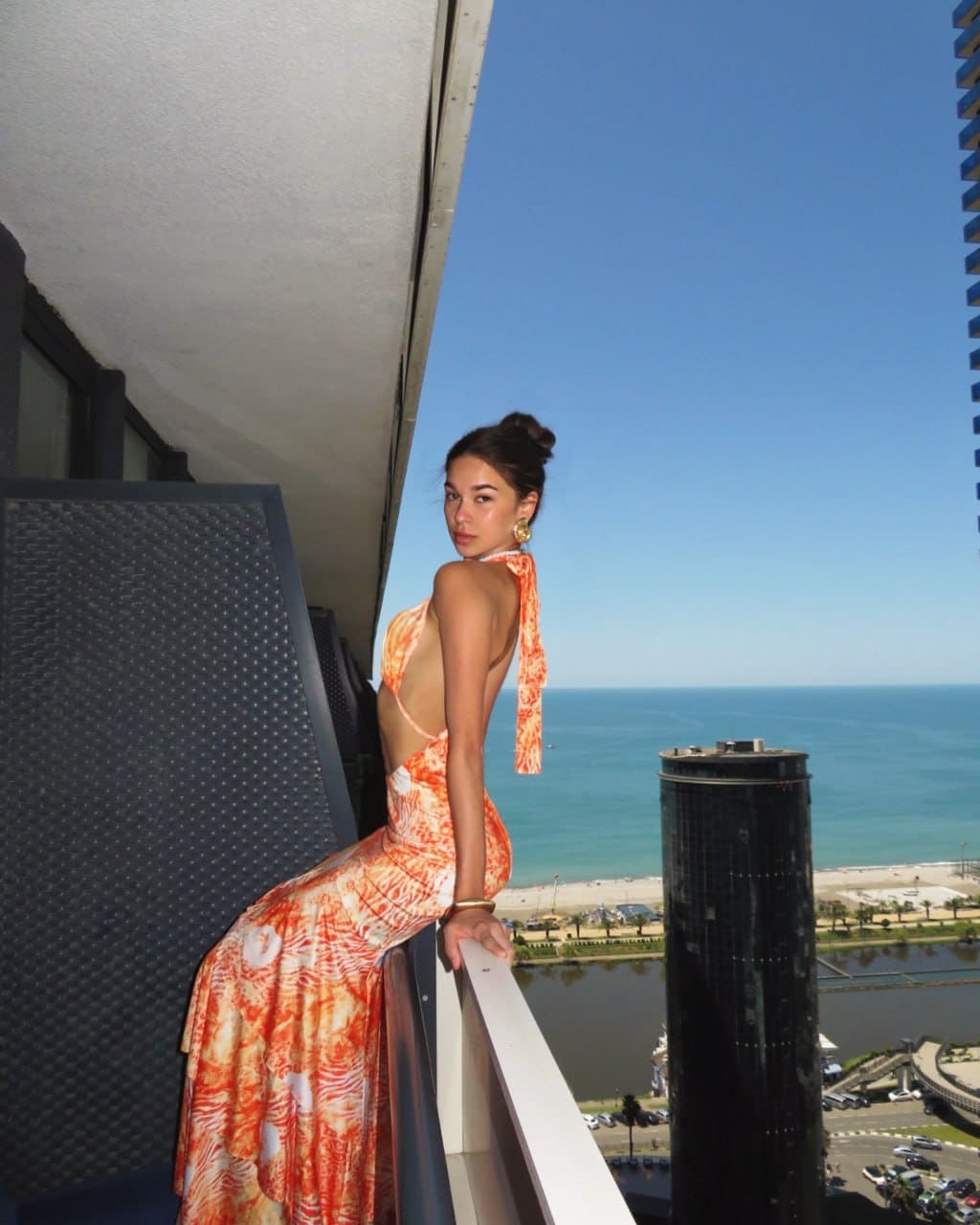 A woman in an orange backless dress stands on a balcony, looking back at the camera with the ocean and city behind her.