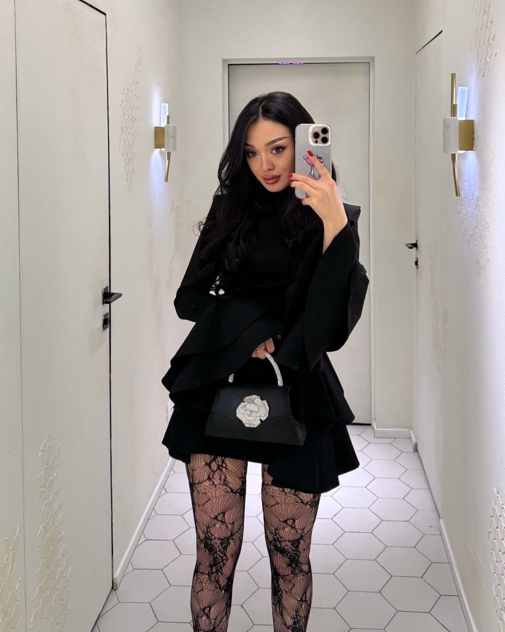 A woman takes a mirror selfie in a white-tiled hallway, wearing a black ruffled dress, lace tights, and holding a floral-embellished bag.