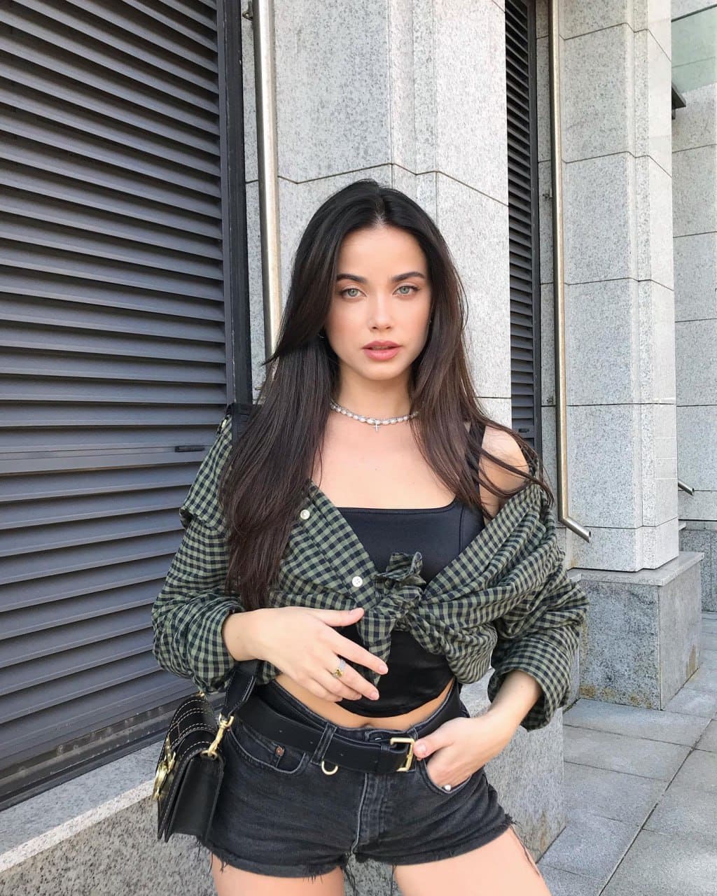 Marina stands outdoors by a stone building in a black crop top, checkered shirt tied at waist, and black denim shorts with one hand on her hip.