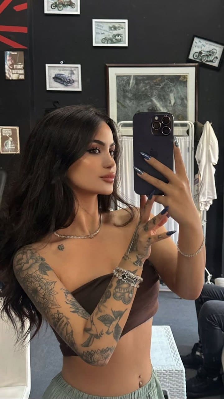 Mirror selfie in a room with framed pictures, wearing a brown strapless tube top and light pants, showing arm tattoos.