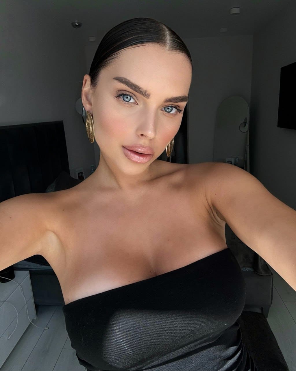 Jean wears a black strapless top and gold hoops, posing in a dim room with a bed and mirror.