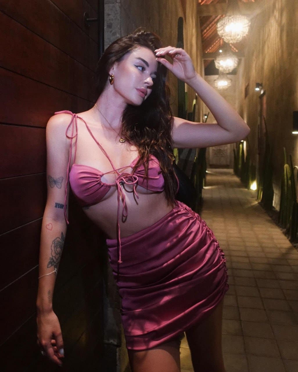 A woman in a pink tie-front bralette and ruched satin mini skirt stands in a narrow outdoor corridor, hand to her forehead.
