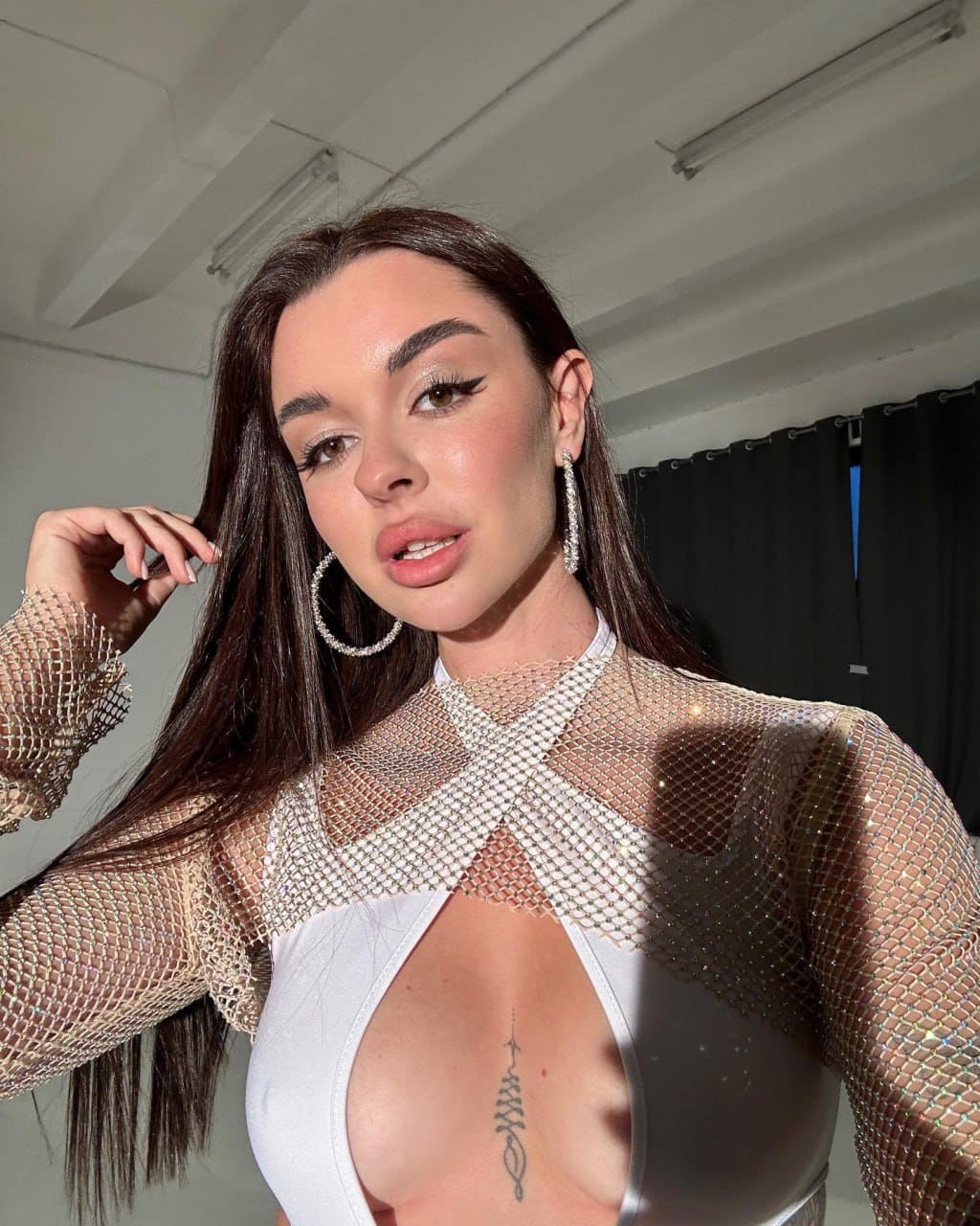 Lavanda poses indoors in a white bodysuit and sheer beaded mesh top, hand near her hair, with a small chest tattoo visible.