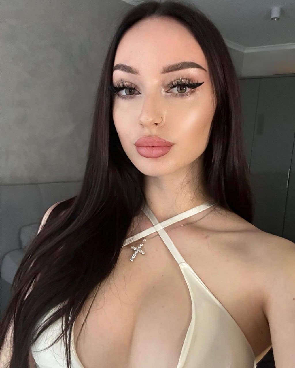 Janine with long dark hair and a nose ring poses in a cream halter top against a plain gray wall.