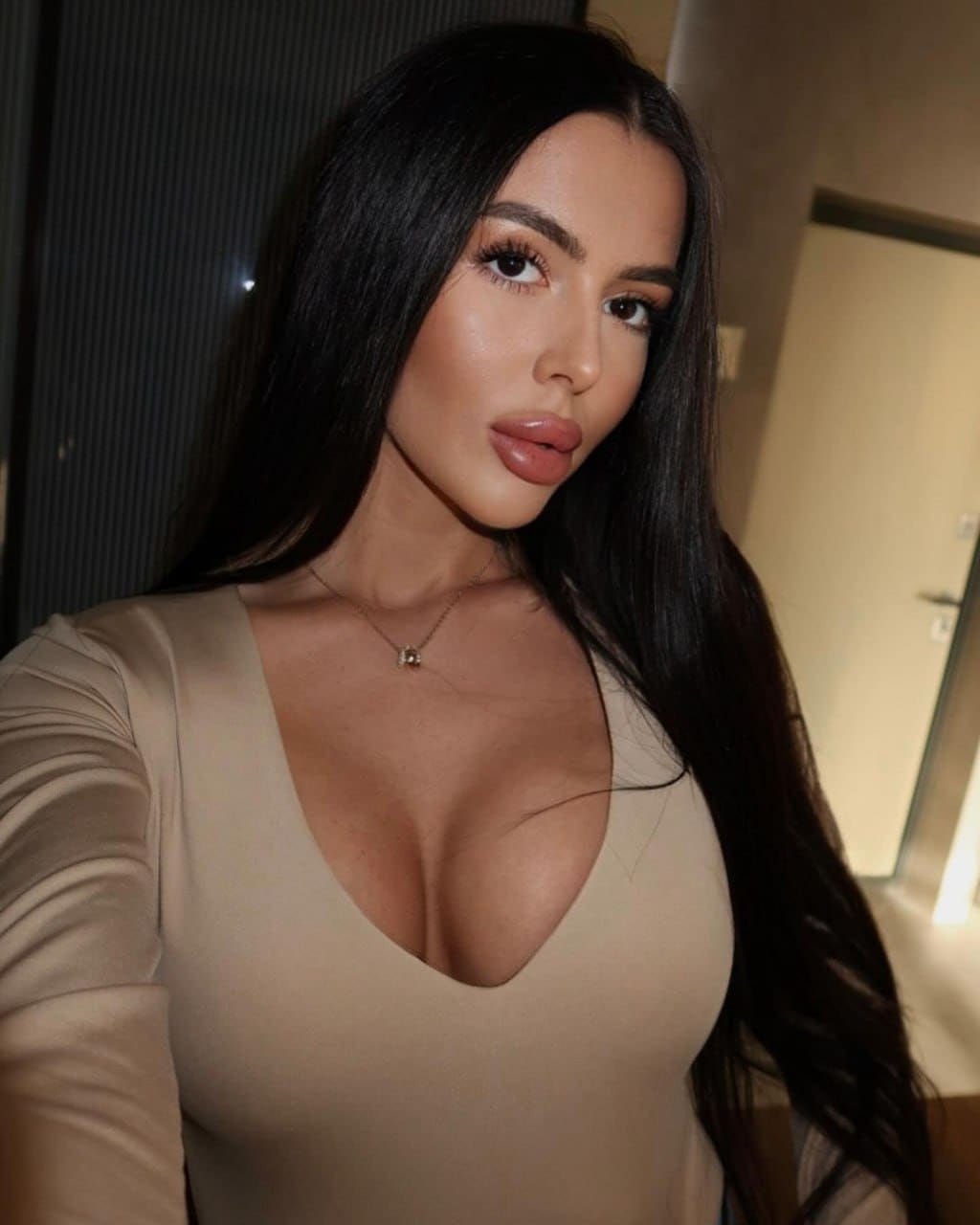Indoors, she takes a selfie in a beige long-sleeved top with a deep V-neck and thin necklace, with a door and dark wall visible behind her.