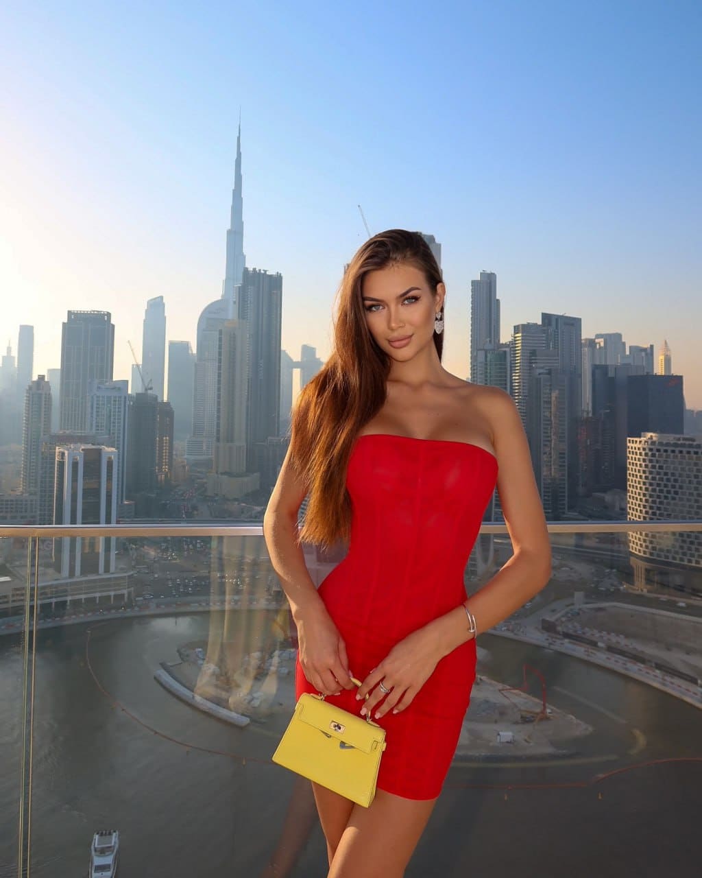 A woman in a red strapless mini dress holds a yellow bag on a rooftop terrace, with a city skyline in the background.
