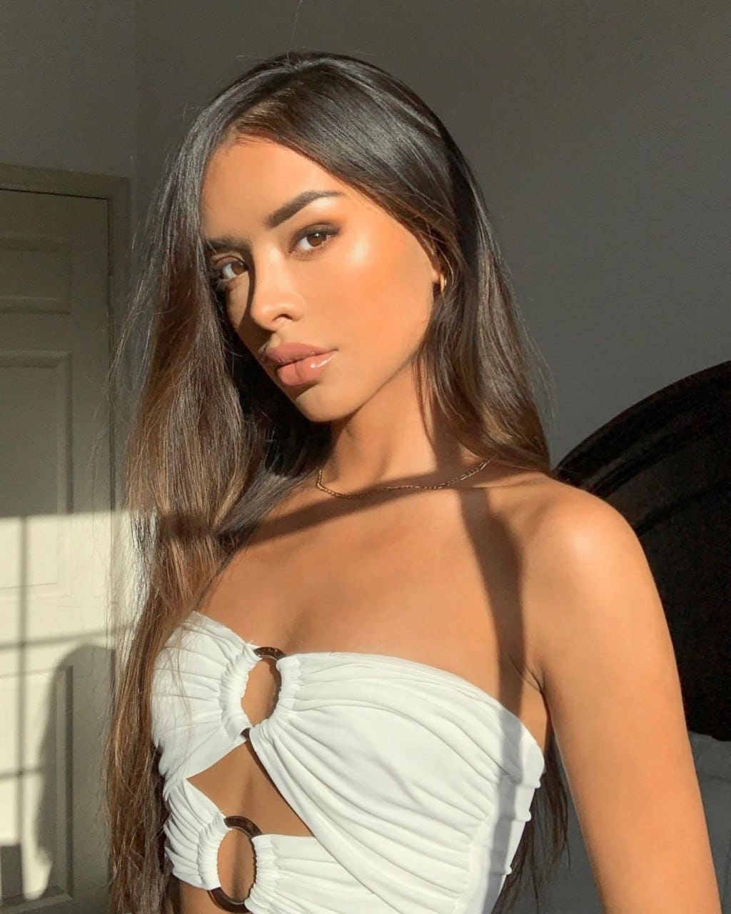 A woman in a white strapless top with cut-outs stands in a sunlit room, looking at the camera.