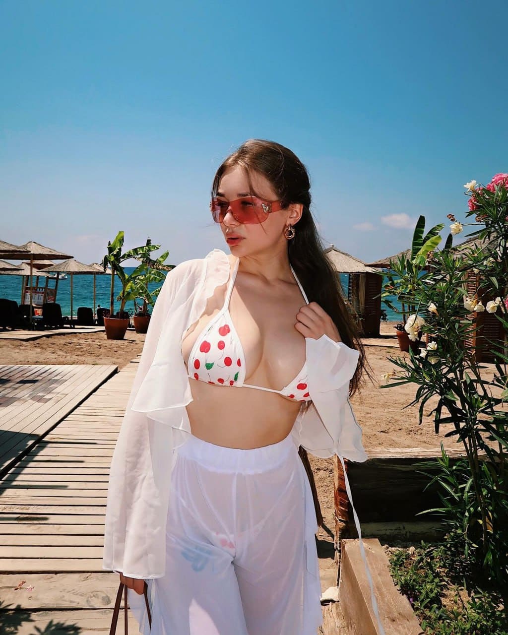 A woman stands on a wooden boardwalk wearing a cherry-print bikini, sheer pants, an open white shirt, and red sunglasses, with umbrellas and sea behind.