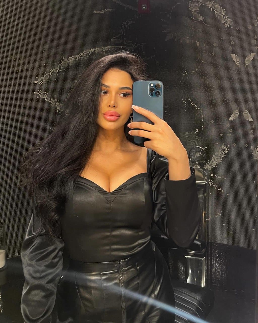 A woman in a black satin top and leather skirt takes a mirror selfie with a blue smartphone against a dark patterned wall.