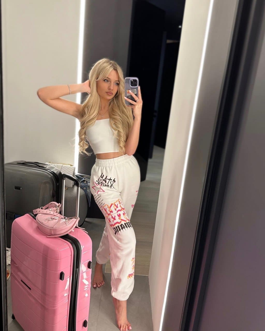 A mirror selfie shows a blonde woman in a white crop top and graphic sweatpants, standing barefoot next to pink and grey suitcases.