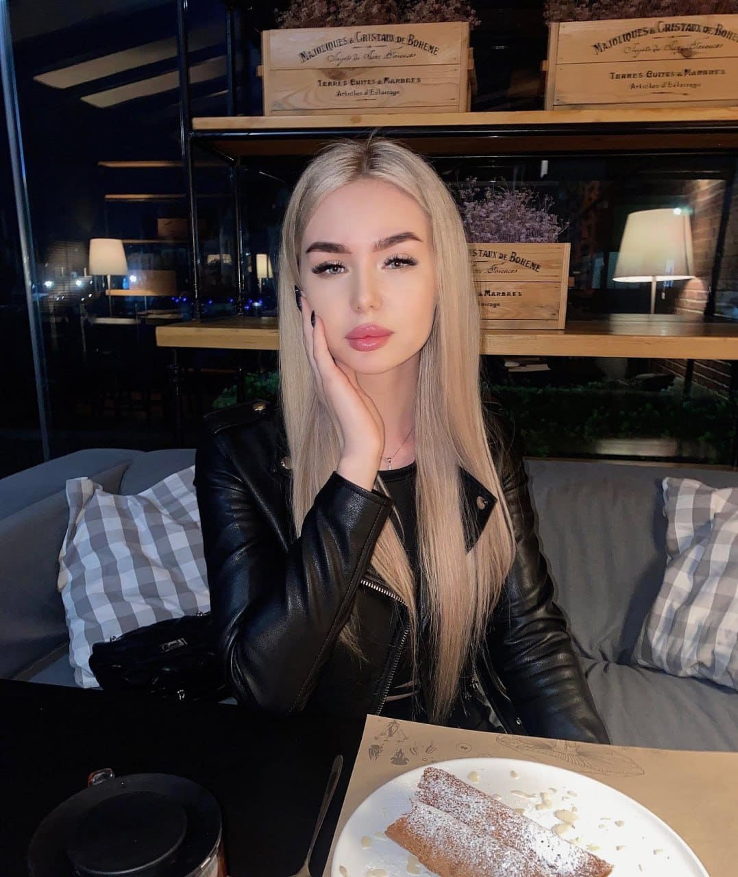 A woman in a black leather jacket sits at a dimly lit table with a plate of powdered pastries, resting her head on her hand.