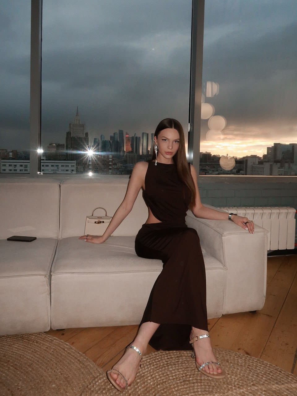 A woman sits on a white sofa before large windows showing a city skyline at dusk, wearing a dark brown dress with a side cutout.