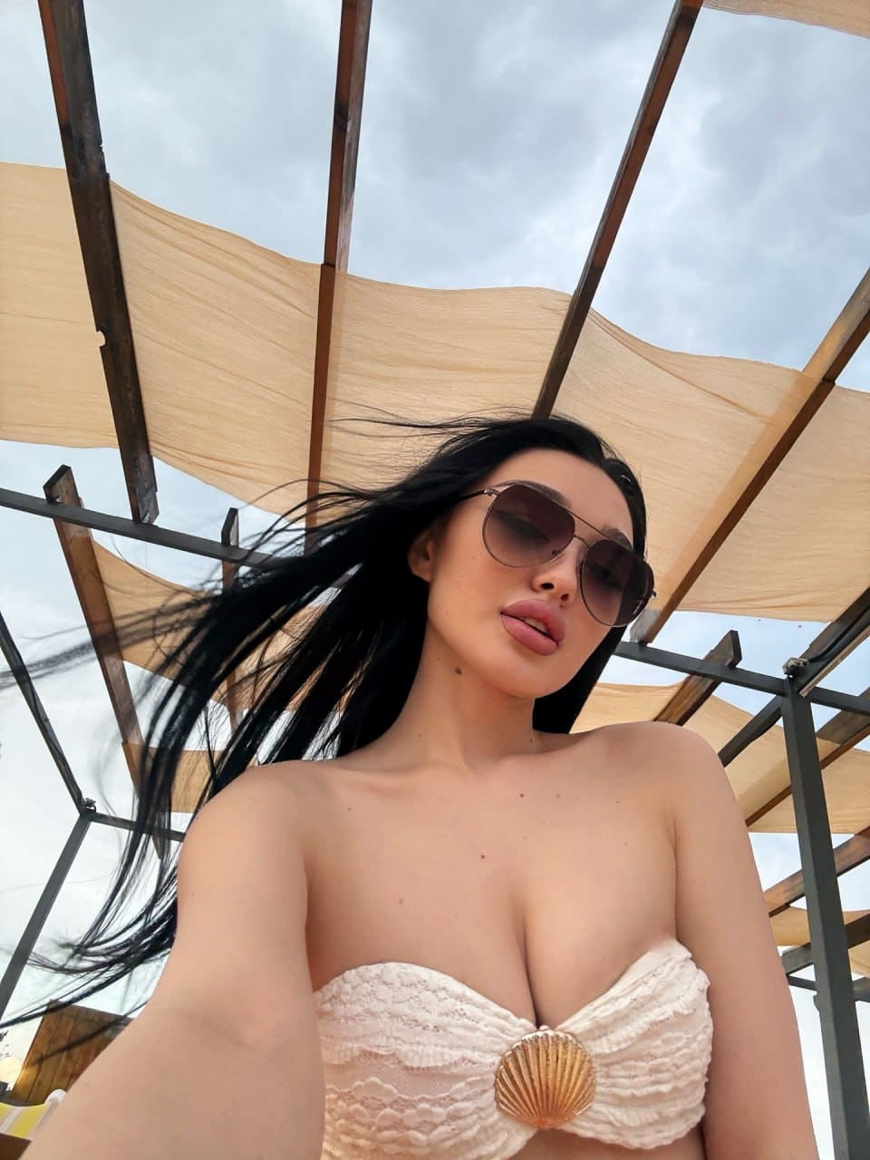 A low-angle selfie captures a woman in sunglasses and a cream strapless lace top, standing under a wooden pergola against a cloudy sky.