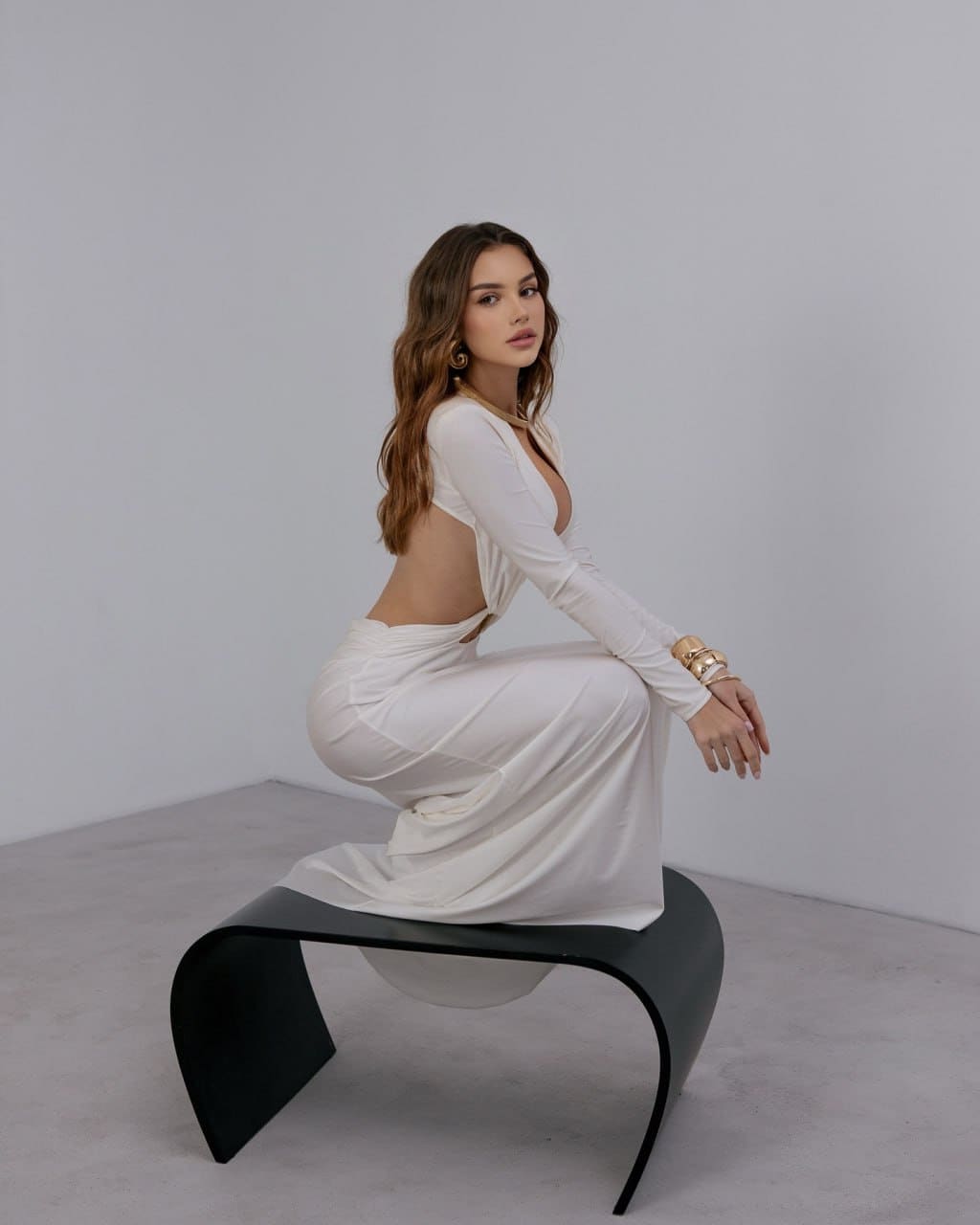 Crouching on a black bench, a woman wears a white long-sleeved dress with a deep front neckline and open back in a gray studio.