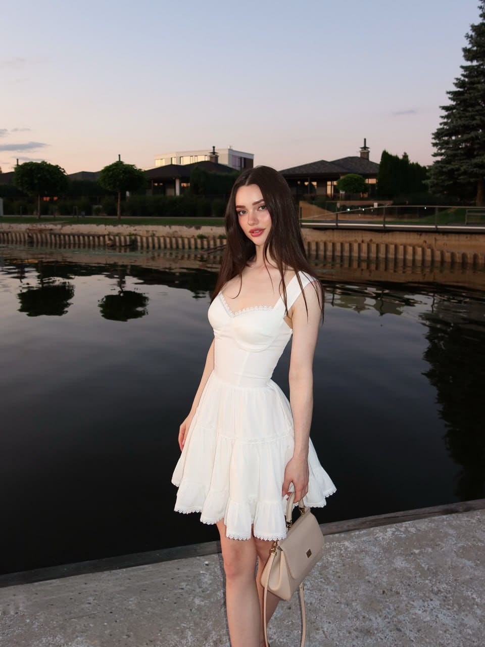 Standing on a concrete ledge at dusk in a white lace-trimmed dress, holding a beige bag near calm water.