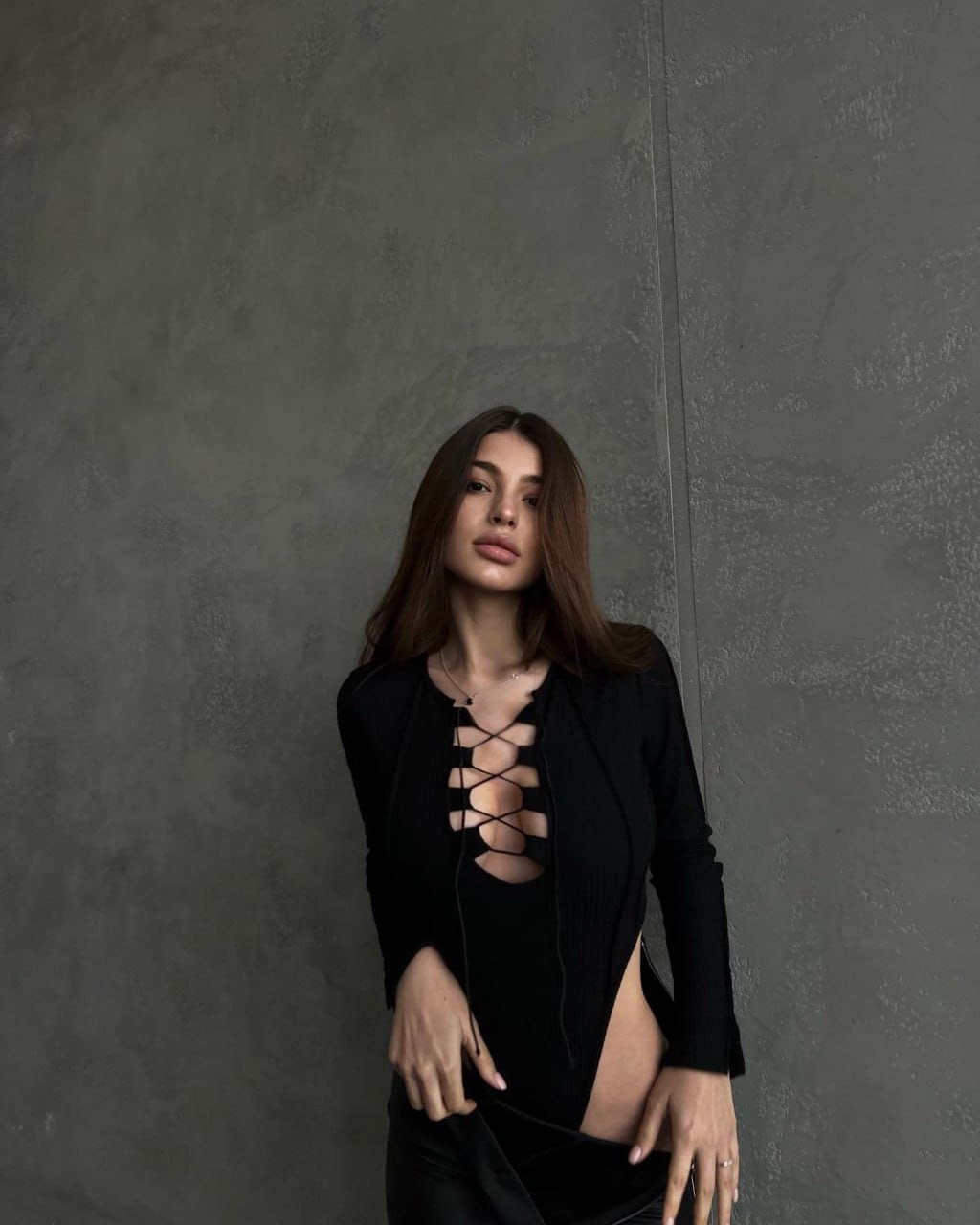 A woman stands against a gray concrete wall wearing a black lace-up top with a side slit and black pants.