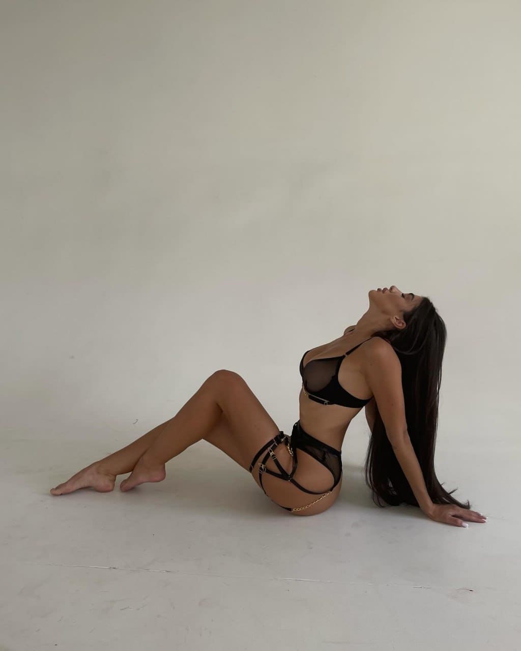 A woman sits on a light floor against a plain wall in black sheer lingerie with gold chains, head tilted back.