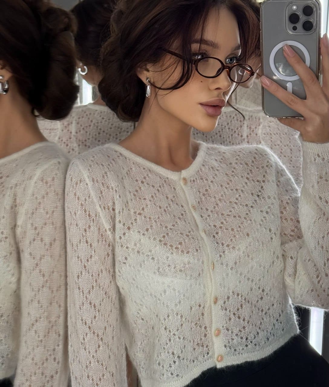 A woman with short dark hair and glasses takes a mirror selfie in a white open-knit cardigan over a dark top, holding a phone.