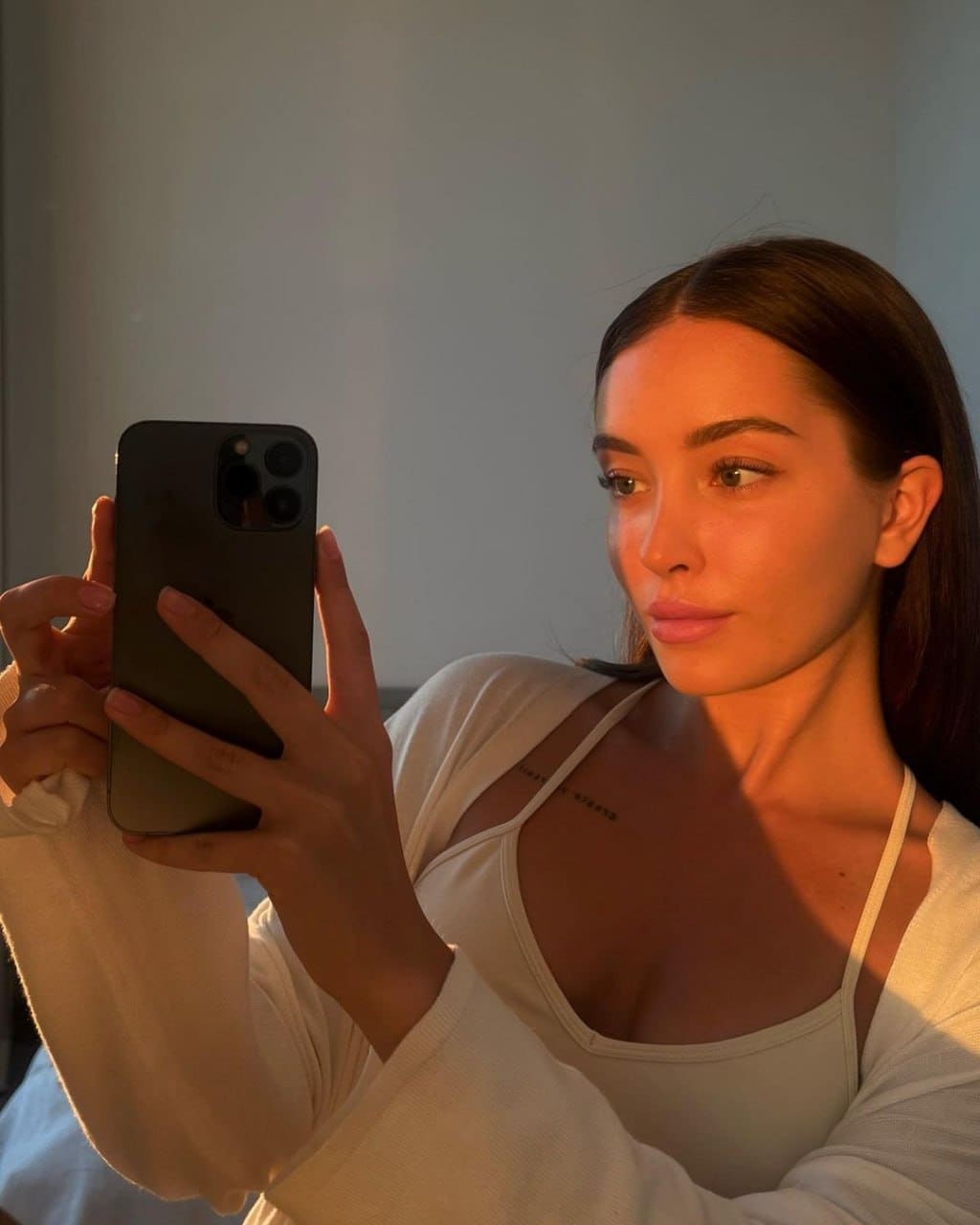 A mirror selfie shows a woman in a white camisole and open cardigan, holding a phone in warm light.