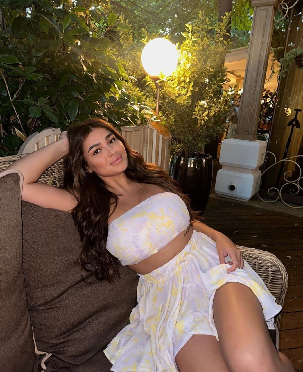 Woman in white and yellow tie-dye outfit sits on wicker chair, arm behind head, near glowing lamp.