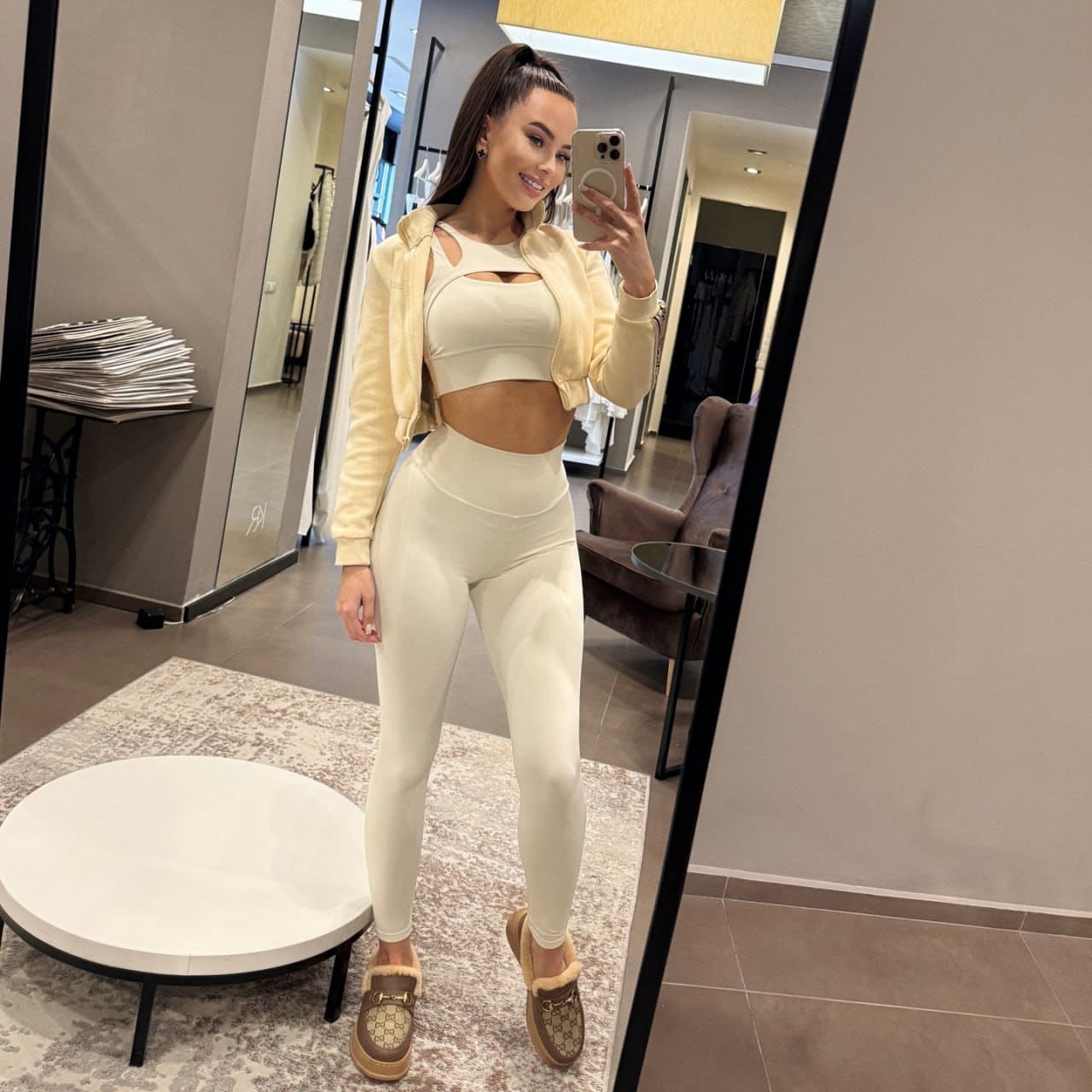 A woman in a cream cut-out outfit and fur-trimmed loafers takes a mirror selfie in a fitting room.