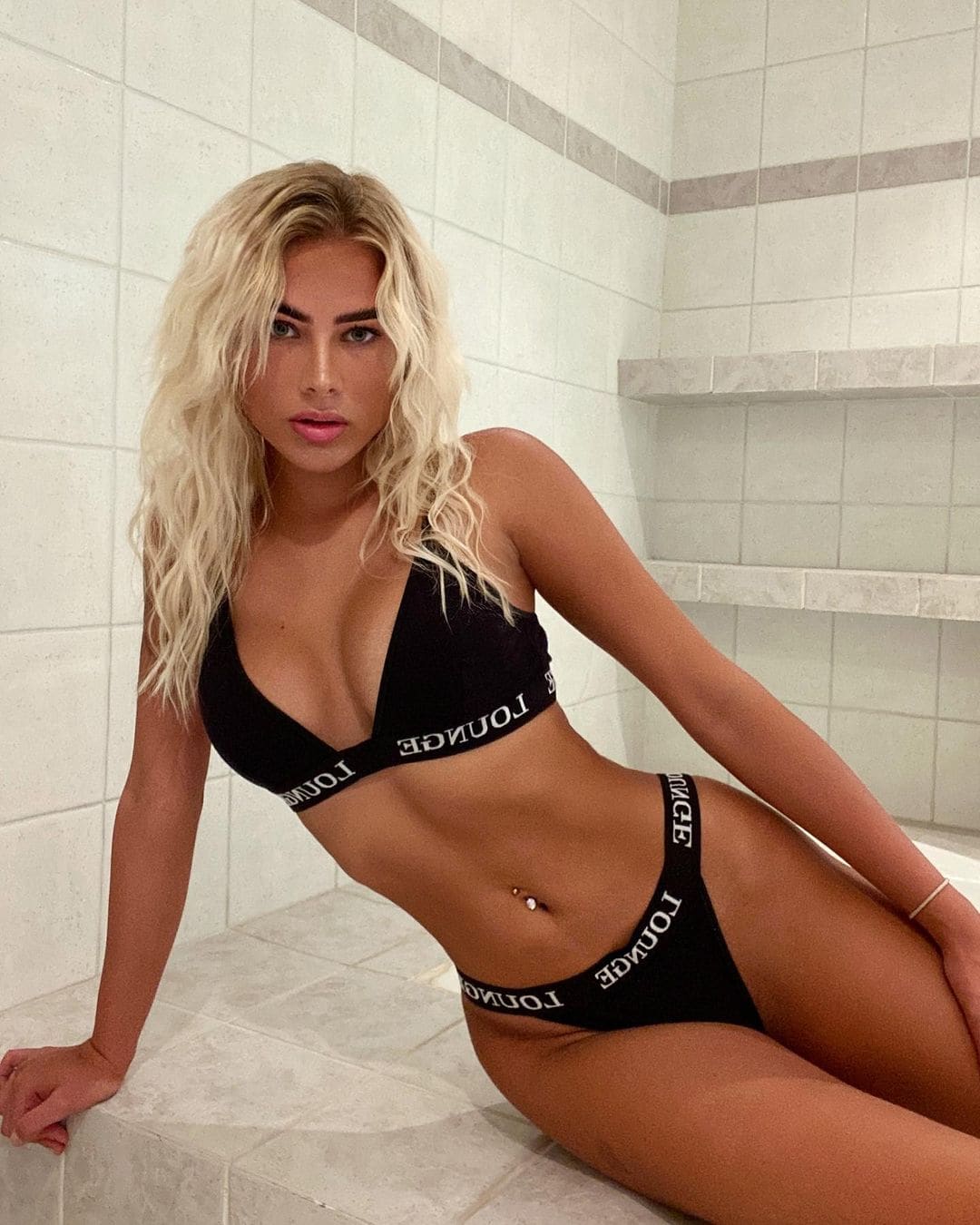 Seated on a tiled ledge in a tiled room, she wears a black bikini with white lettering, leaning forward with one hand on the surface.