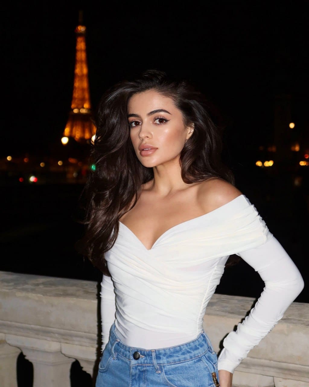 A woman in a white off-shoulder top and jeans stands at night by a stone balustrade, hand on hip, near a lit tower.