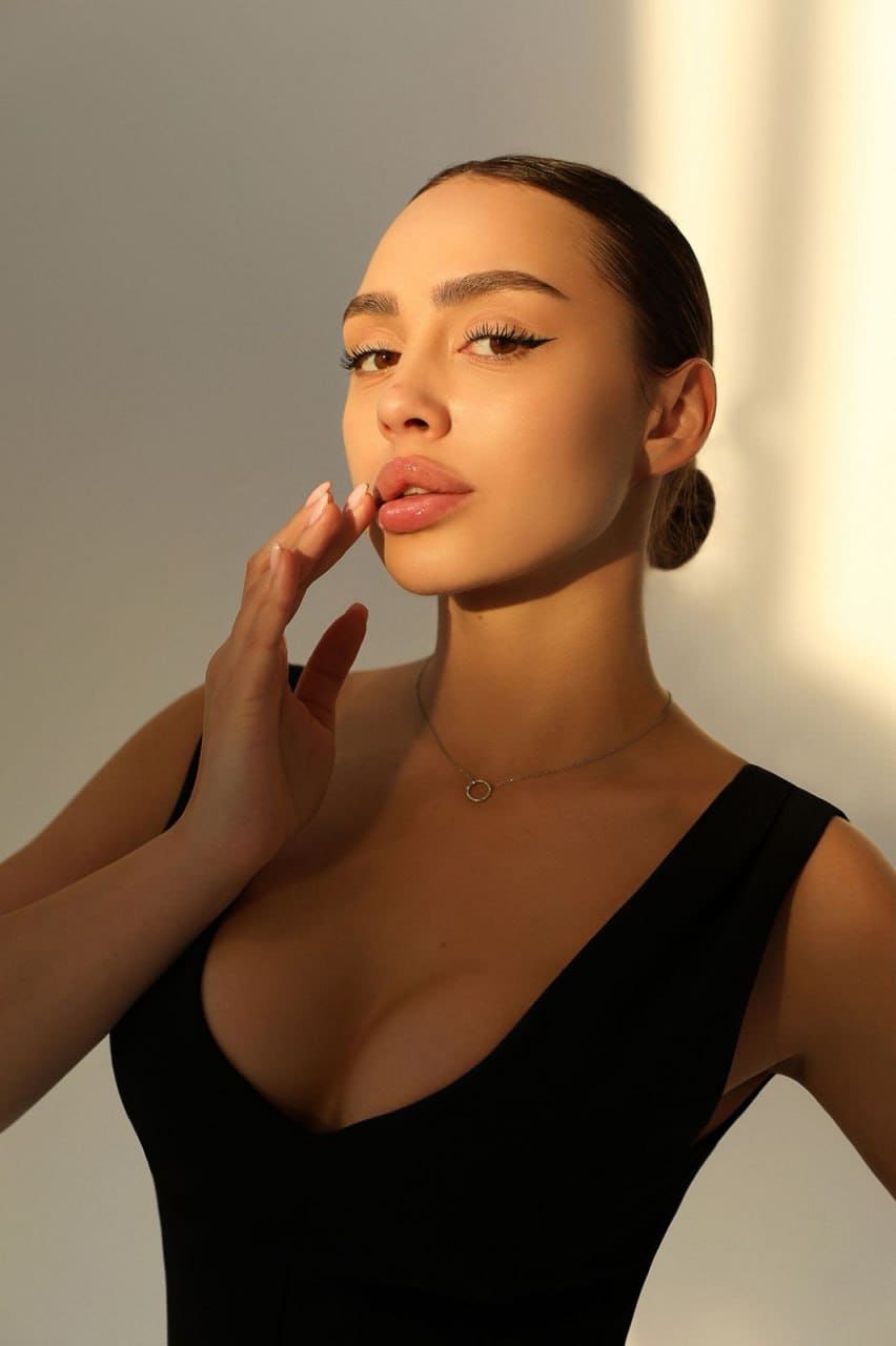 Mariana with dark hair in a low bun wears a black V-neck top, hand to lips, against a lit wall.