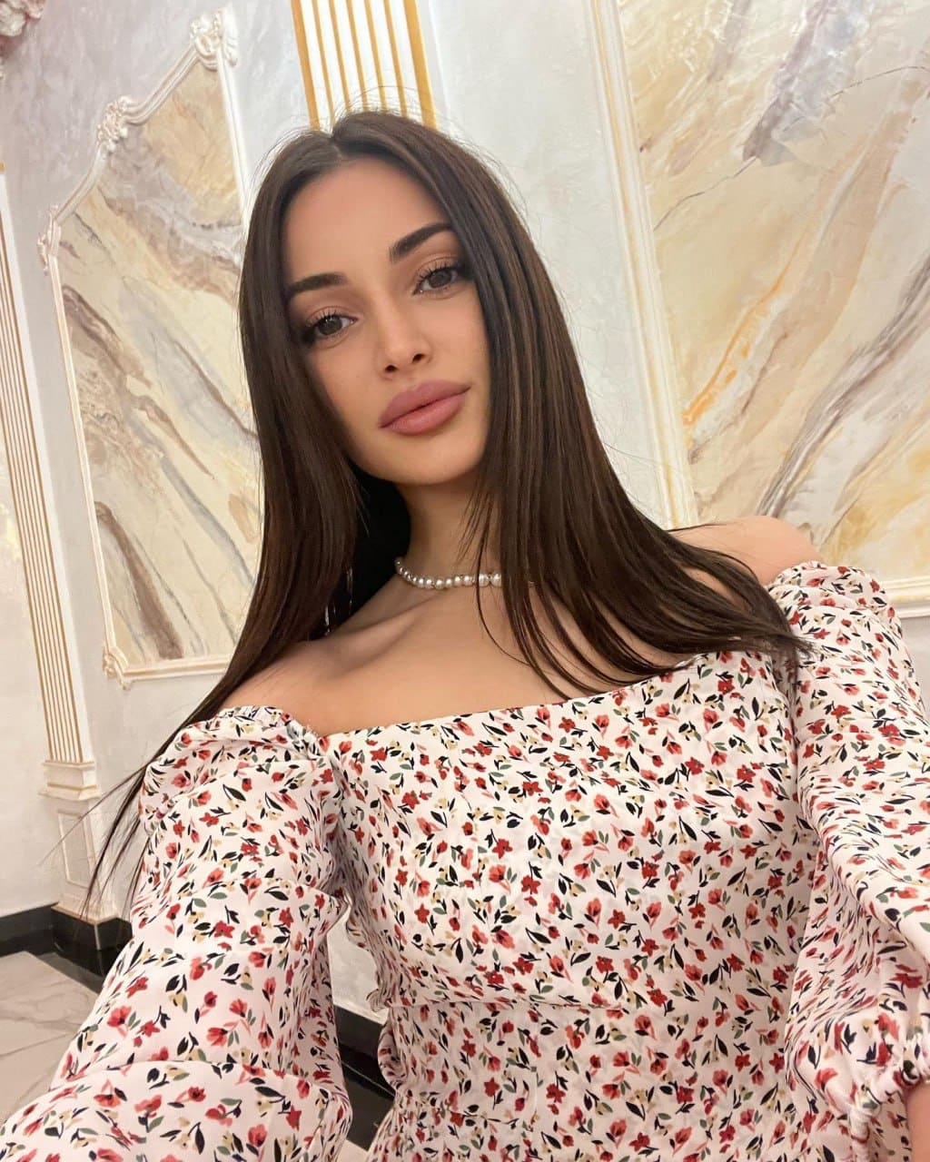 A woman in an off-the-shoulder floral dress and pearl necklace stands before a marble wall with gold trim.