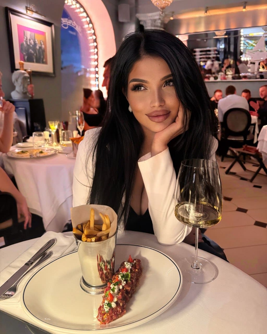A woman sits at a restaurant table resting her chin on her hand, wearing a white blazer over a black top, with food and white wine in front.