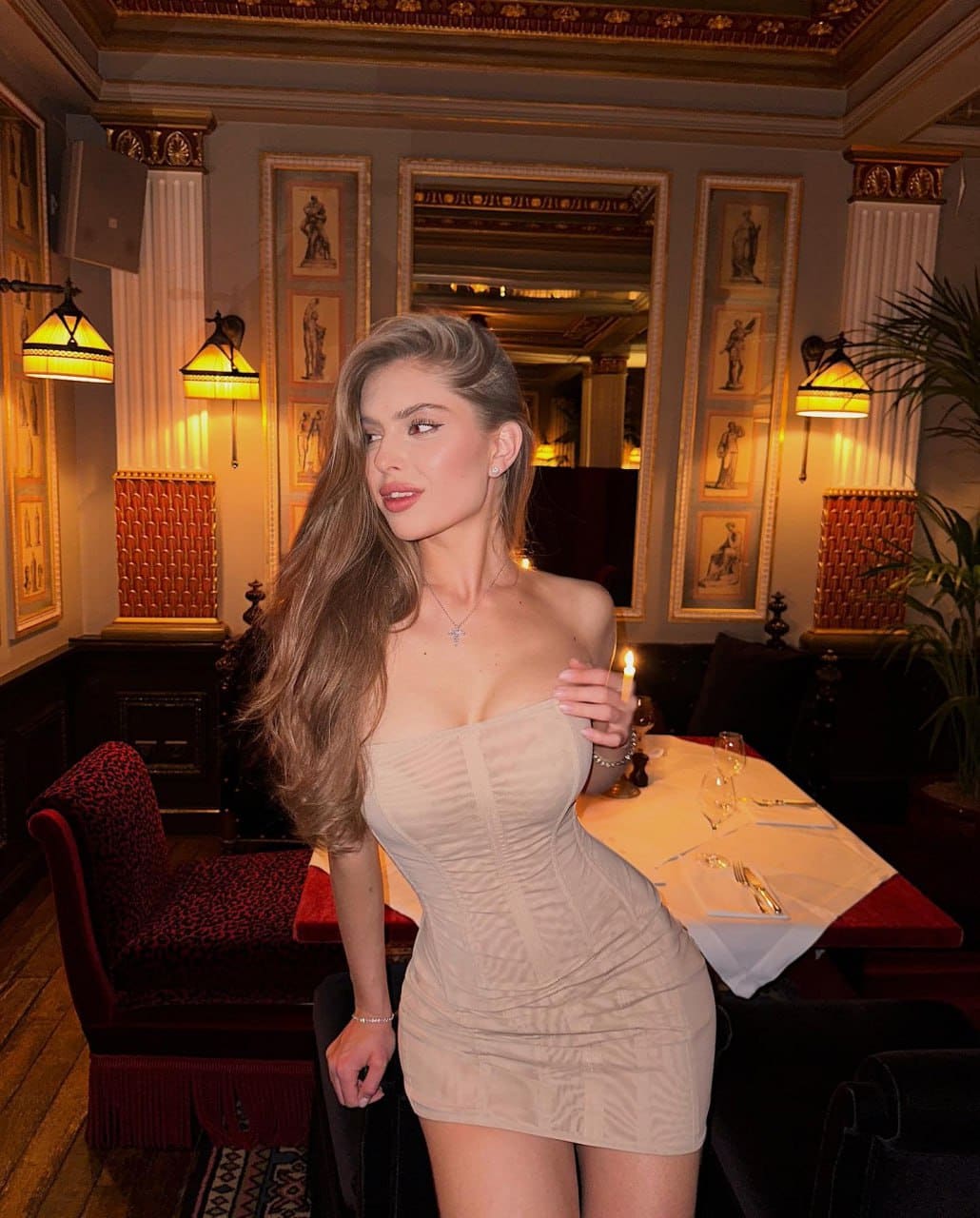 Woman in a strapless beige mini dress stands in a dim restaurant, holding a lit candle near her chest.
