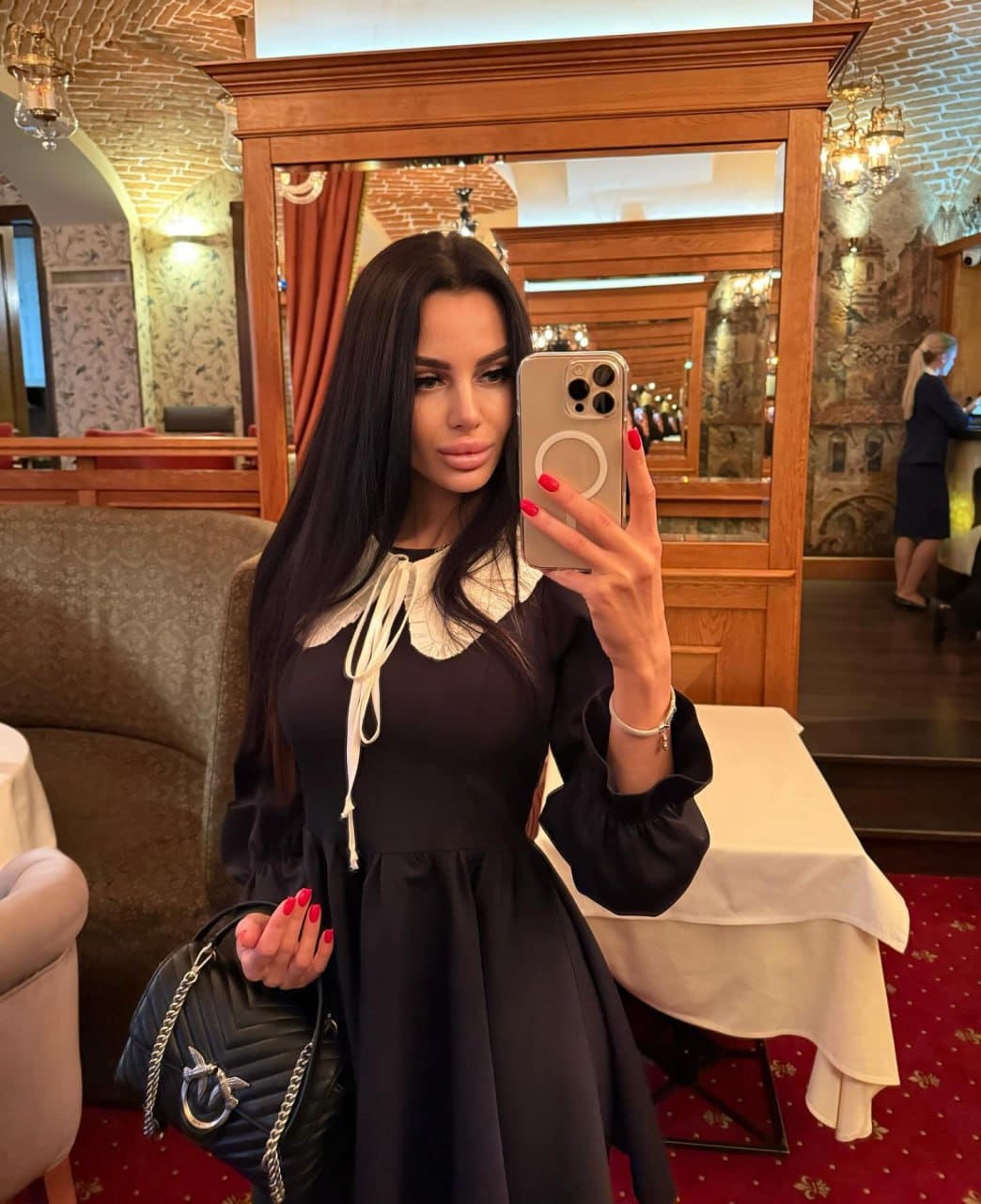 Madelina takes a mirror selfie in a restaurant, wearing a black dress with a white lace collar and holding a black quilted handbag.
