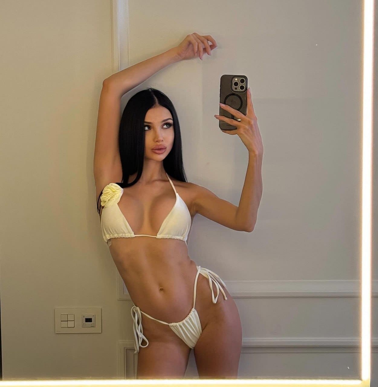 A woman takes a mirror selfie in a white ruffled bikini, one arm raised, standing in front of a lighted mirror frame.
