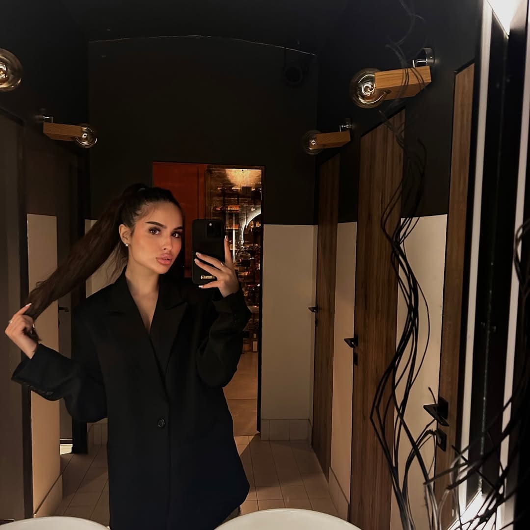 A woman in a black blazer takes a mirror selfie in a bathroom with wooden stalls and exposed bulbs, touching her ponytail.