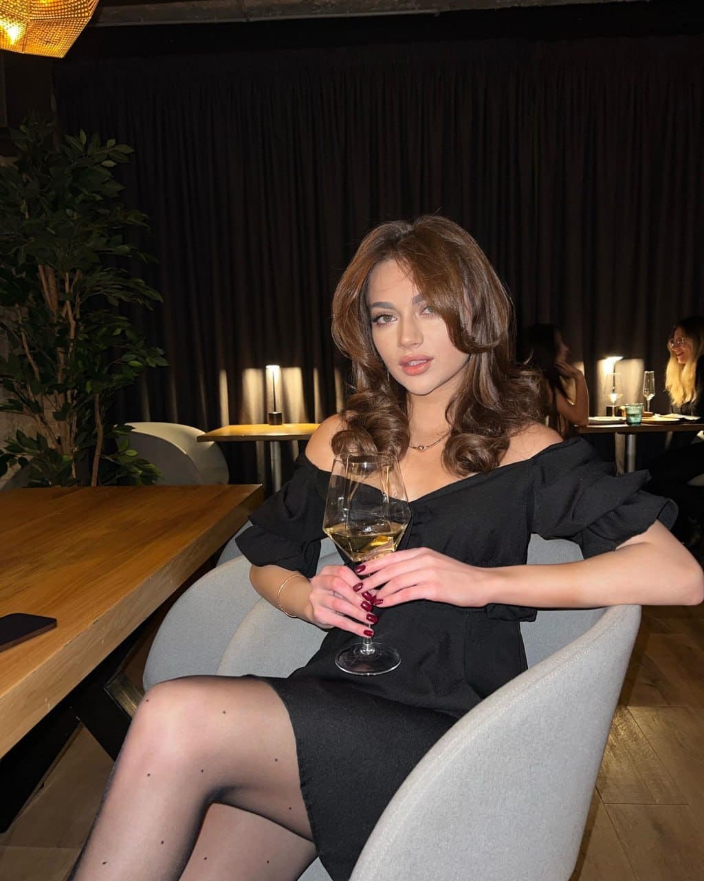 A woman in a black off-shoulder dress sits in a grey armchair holding white wine, against dark curtains and plants.
