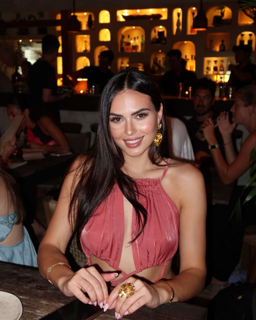 A woman in a dusty-pink halter top sits at a wooden table in a dimly lit restaurant, hands resting on the surface.