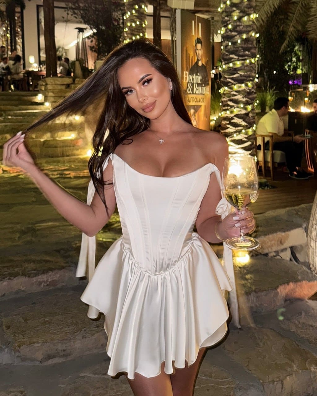 A woman in a white off-shoulder dress holds wine outdoors at night, with string lights and trees behind her.