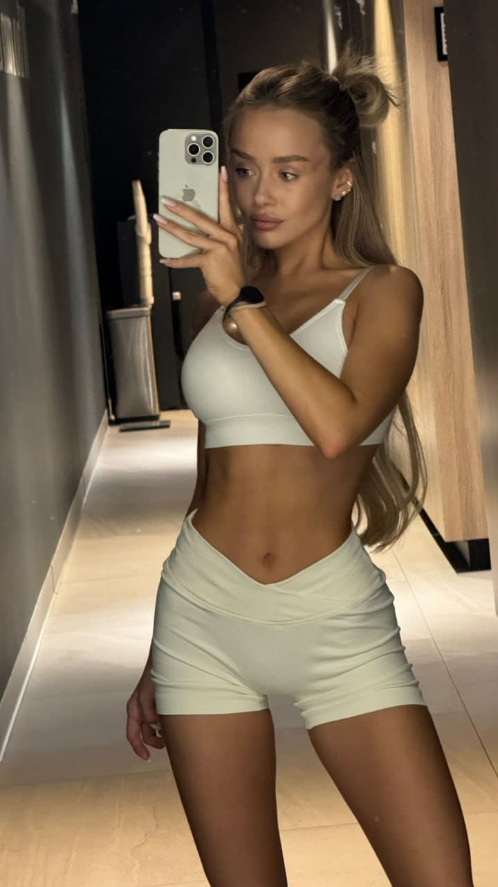 Mirror selfie in a hallway showing a white sports bra and high-waisted shorts, hair in a high bun.