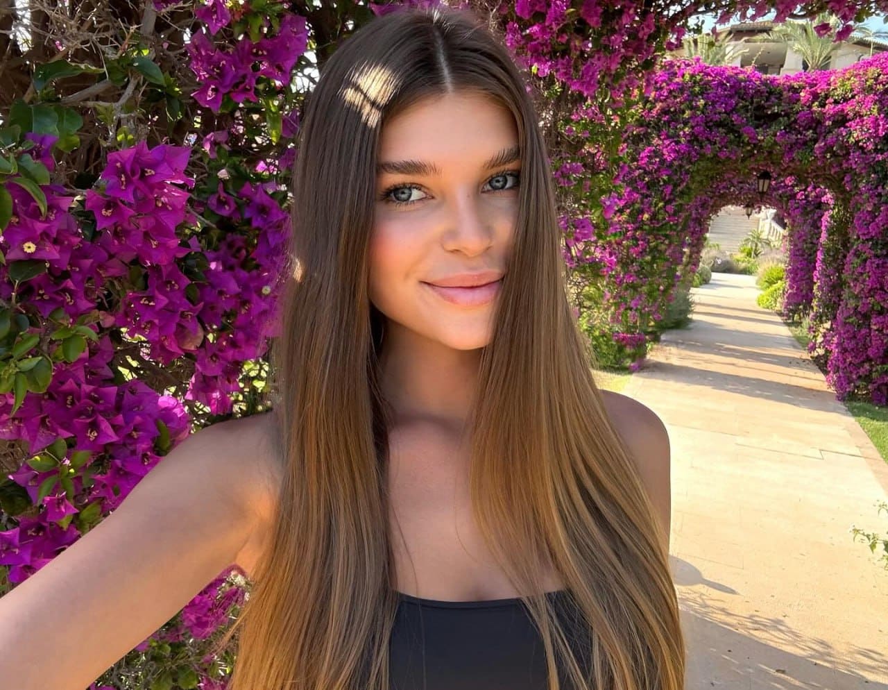 Giulia stands outdoors in a black strapless top, smiling slightly before a path lined with purple bougainvillea flowers.