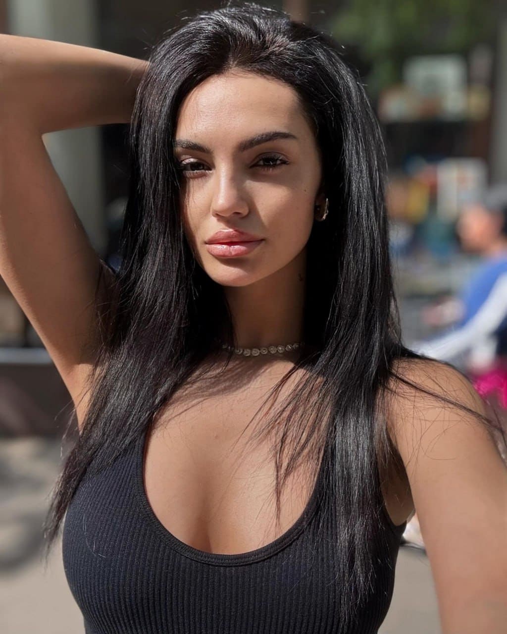 Karina poses outdoors in a black ribbed tank top with a deep scoop neckline, one arm raised behind her head, against a blurred street background.