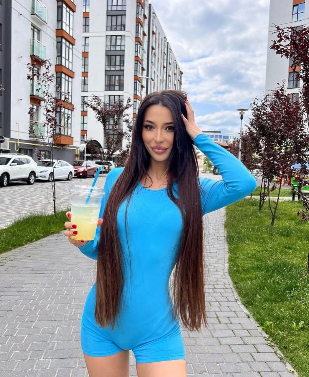 Standing on a paved walkway, she wears a blue romper and holds a drink with a blue straw.