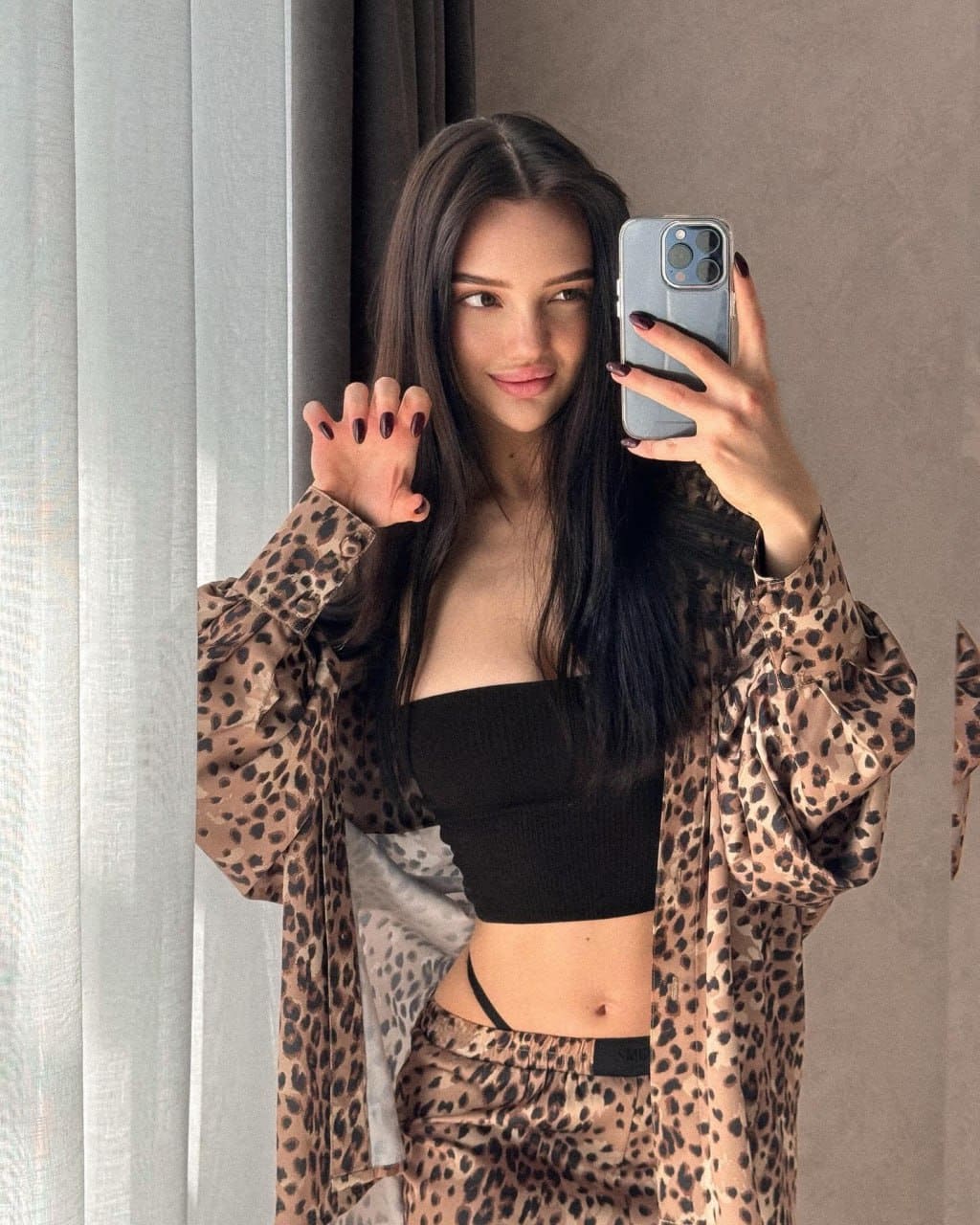 Mirror selfie of a woman in an open leopard-print shirt over a black crop top, standing by a window with sheer curtains.