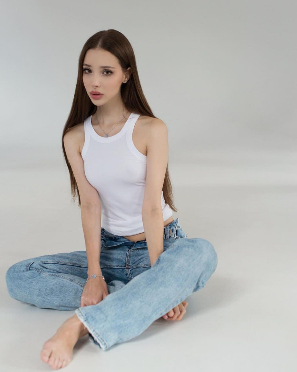 A woman sits cross-legged on a gray studio floor in a white tank top and blue jeans, barefoot, wearing a thin necklace and bracelet.