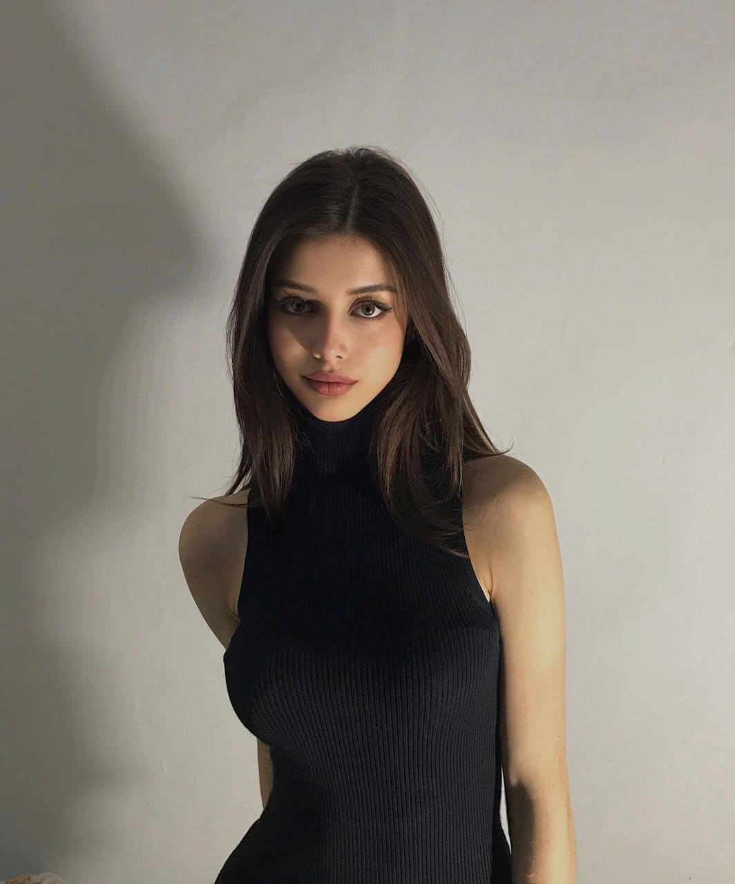Woman with long dark hair faces the camera in a sleeveless black ribbed turtleneck, posed against a plain light wall.