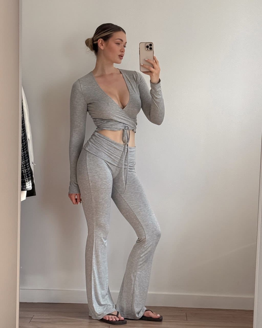 A woman with a low bun wears a grey long-sleeved wrap top and flared pants, taking a mirror selfie on a wooden floor against a white wall.