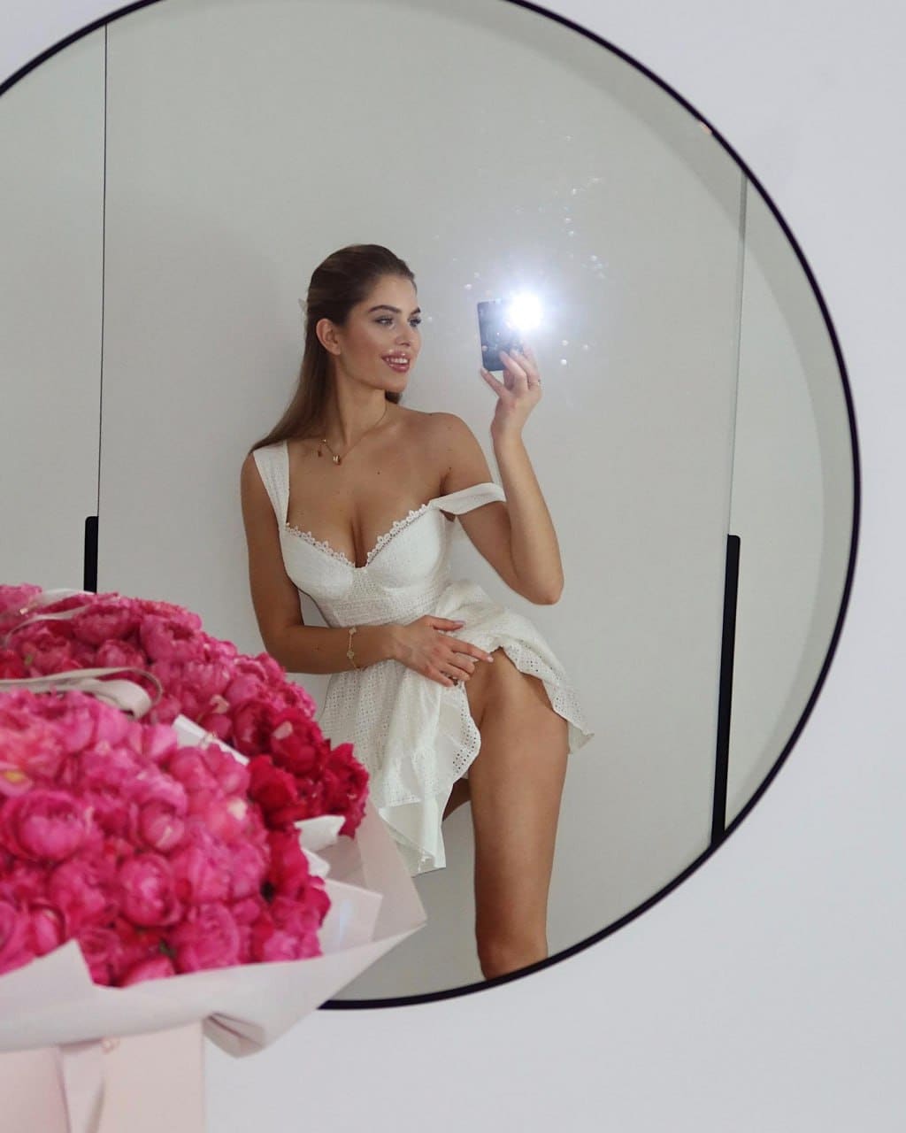 Mirror selfie in a white off-shoulder lace dress, holding a phone with flash, pink flowers in foreground.